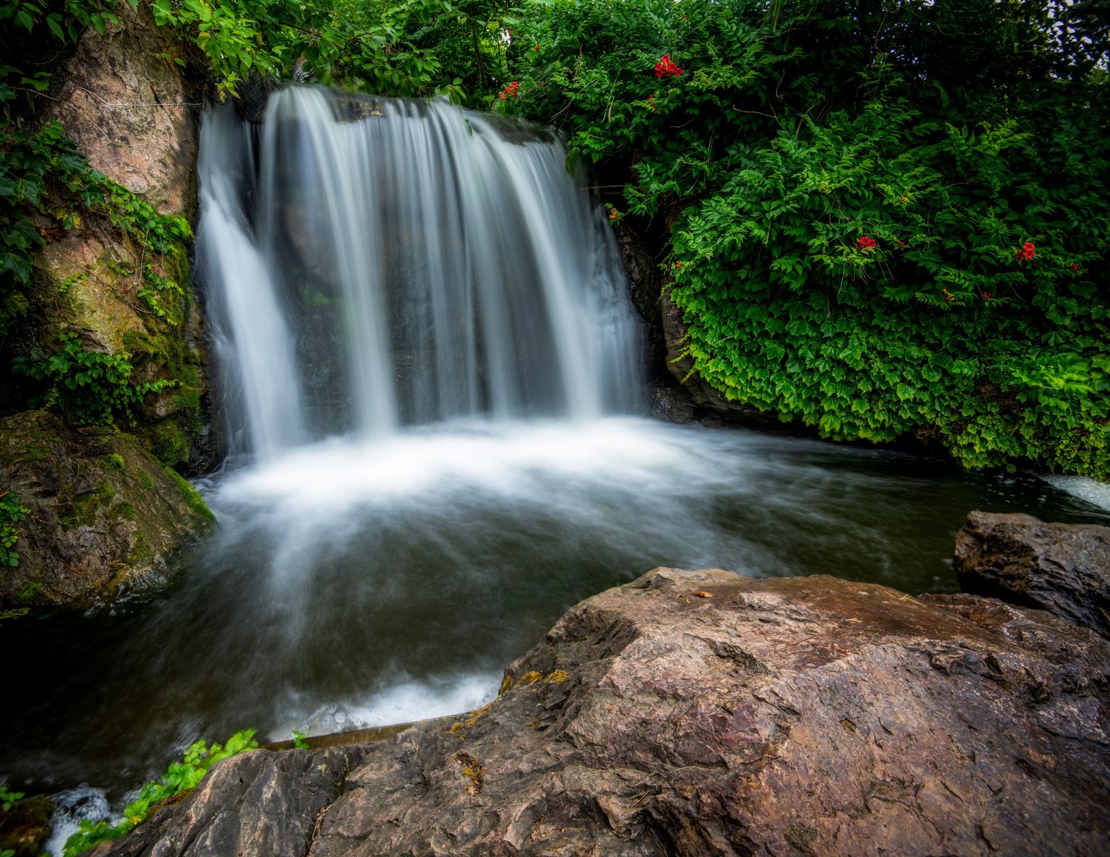 A wide waterfall spilling over rock ledges into a clear pool surrounded by greenery